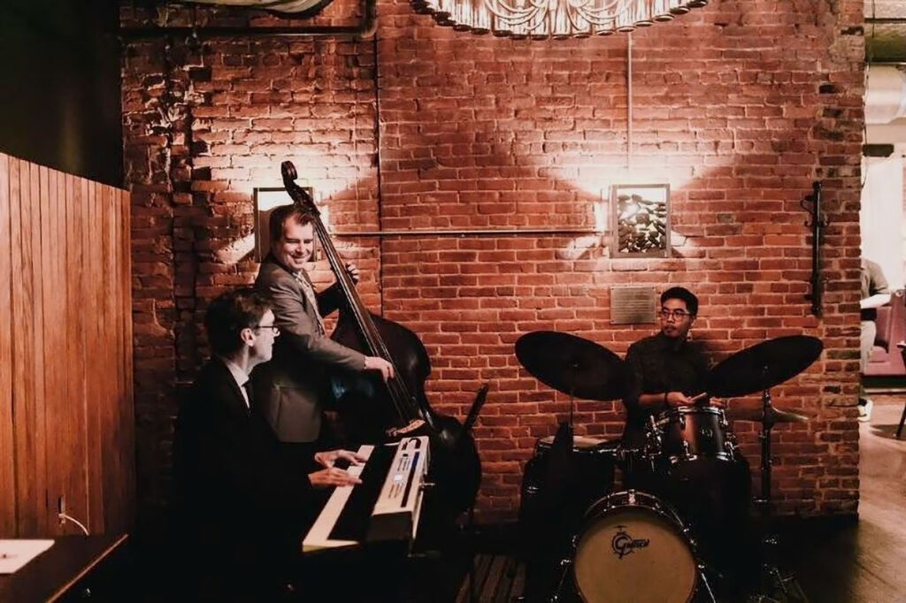 A jazz trio (keyboard, upright bass, and drums) playing against a brick wall background