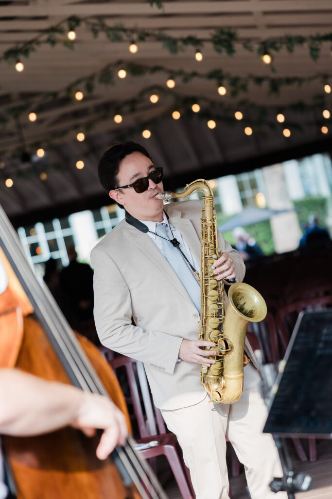 A musician in sunglasses and light-colored suit playing saxophone. Bass and bassist's arm visible in foreground.